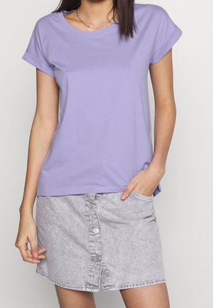 Kvinde iført en lavendel kortærmet t-shirt og en lysegrå denimnederdel med knapper foran, står med den ene hånd i lommen.