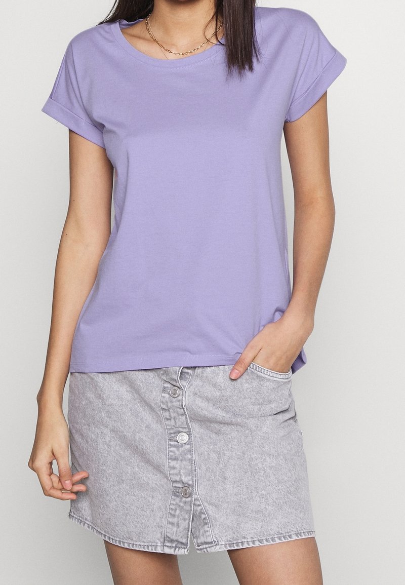 Femme portant un t-shirt lavande à manches courtes et une jupe en jean gris clair à boutons sur le devant, debout avec une main dans la poche.