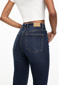 Jeans in denim blu scuro con vita alta, caratterizzati da due tasche posteriori e una patch in pelle in vita. Tessuto liscio, design a gamba dritta.