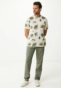 Kurzärmliges Hemd mit crèmefarbenem Grund und grünem Blättermuster; kombiniert mit olivgrünen Hosen und weißen Sneakers.
