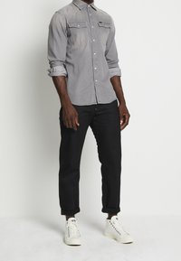 Camicia di jeans grigia con due tasche sul petto, colletto a punta, maniche arrotolate, indossata con pantaloni neri e sneakers bianche.
