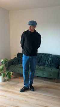 Zwarte jas, blauwe spijkerbroek en zwarte schoenen. Lichtblauwe pet. Staand in een kamer met houten vloer en een groene bank met gestructureerde kussens.