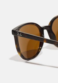 Occhiali da sole tortoiseshell marroni con montatura rotonda e lenti marroni. Presenta un logo Ray-Ban argentato sulla stanga. Finitura liscia e lucida.