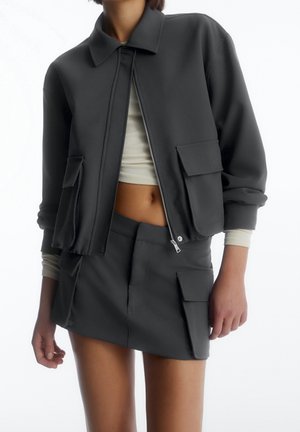 Femme portant une veste courte zippée gris foncé avec de grandes poches à rabat et une mini-jupe assortie, sous laquelle elle porte un crop top côtelé crème.