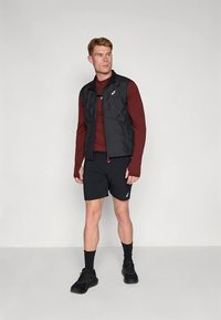 Svart quiltad väst över en mörkröd långärmad tröja, kombinerad med svarta shorts. Stiliserad med svarta sneakers och ankelstrumpor. Minimalistisk design.