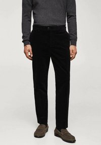 Pantaloni in corduroy neri con righe verticali, dotati di chiusura a bottone e passanti per cintura, abbinati a un top grigio a maniche lunghe e scarpe marroni.