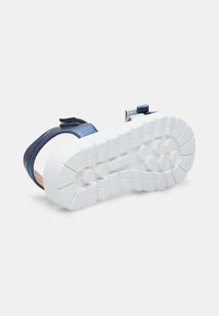 Modra metalna sandal s nastavljivim trakom, bela teksturirana podloga in ravna peta. Ima trajno gumijasto podlago z vzorci za oprijem.
