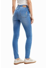 Hubené modré džíny s hladkou texturou, vysokým pasem a prošitými detaily na zadních kapsách. Nosit s bledě zbarvenými teniskami.