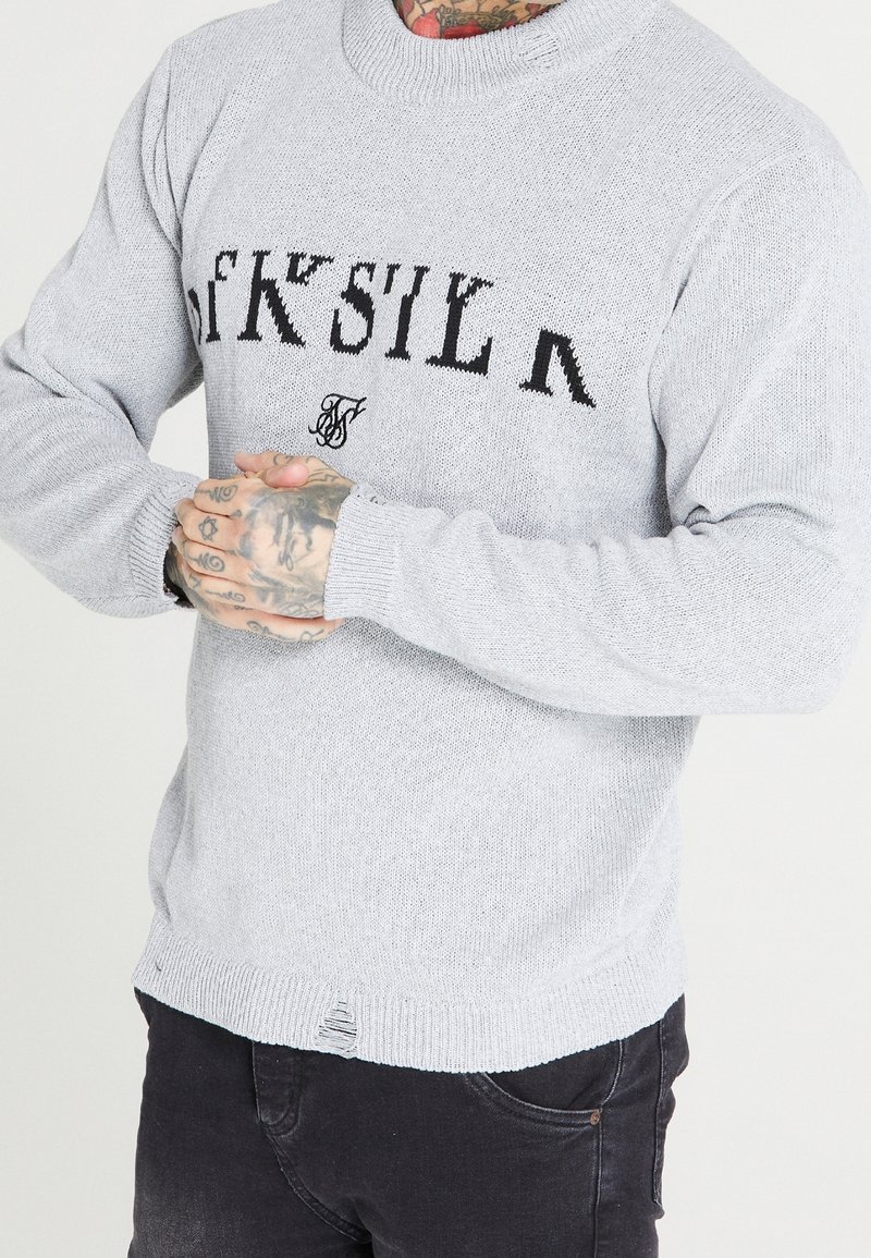 Pull en maille gris clair avec un logo en texte noir, un col rond côtelé et des détails légèrement effilochés au niveau de l'ourlet et des poignets.