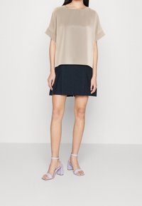 Nainen, jolla on yllään beige lyhythihainen paita, musta laskostettu mini-hame ja lila paksupohjaiset nilkkaremmisaappaat, seisoo yksinkertaisella taustalla.