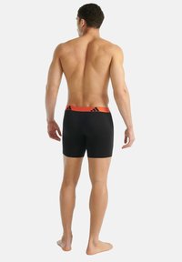 Černé boxer briefs s oranžovým pasem, který má tři černé pruhy. Tkanina vypadá hladce a přiléhavě, bez viditelných log.