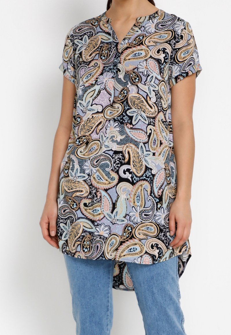 Tunique ample à manches courtes dans un motif paisley noir et pastel, avec un encolure fendue et un ourlet arrondi. Portée avec un jean bleu clair.