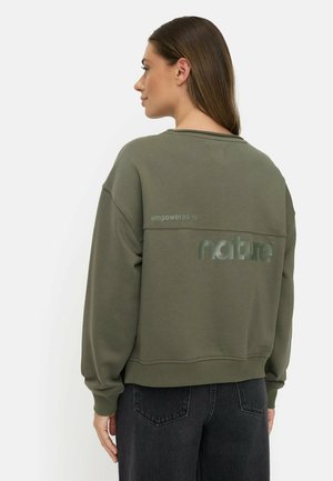 Femme vue de dos portant un sweat-shirt vert olive avec le texte "empowered by nature" dans le dos et un jean noir.