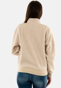 Sudadera de forro polar beige con un cuello alto, superficie texturizada y dobladillo acanalado. Presenta hombros caídos y se combina con unos jeans de mezclilla azules.