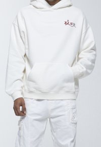 Felpa bianca in misto cotone, con tasca frontale, polsini a costine e un piccolo logo bordeaux sul lato sinistro del petto. Abbinata a pantaloni bianchi.