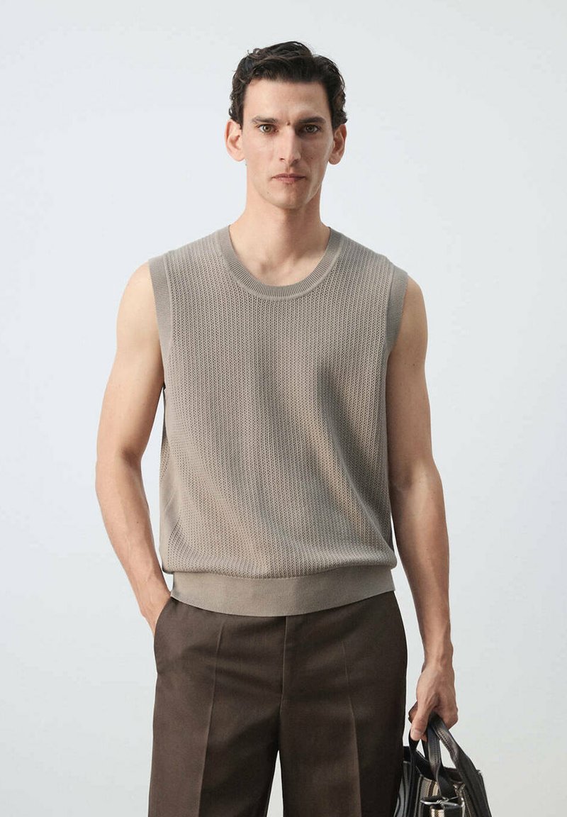 Homme portant un haut en maille beige sans manches et un pantalon marron, tenant un sac en cuir noir avec poignées sur fond uni.