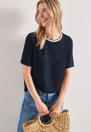 Femme souriante aux cheveux blonds porte un haut bleu marine à manches courtes et un jean, tenant un sac en paille tressée à poignée ronde devant un mur uni.