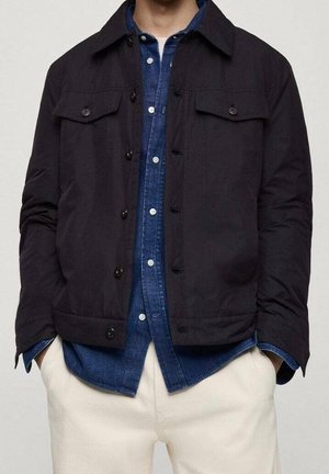 Mann trägt schwarze, geknöpfte Jacke über einem blauen Jeanshemd mit weißen Knöpfen, kombiniert mit cremeweißen Hosen und Händen in den Taschen.