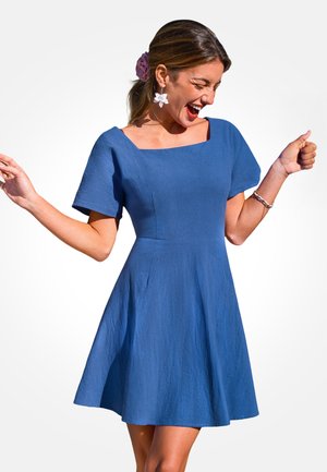 Donna sorridente che balla con gli occhi chiusi, indossa un vestito blu a maniche corte, una forcina per capelli a forma di fiore e orecchini a forma di fiore su sfondo bianco.