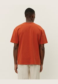 Tricou din bumbac de culoare portocalie ruginit, cu un croi lejer, guler rotund și mâneci scurte, expus din spate. Suprafață simplă, fără modele.