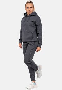 Grauer Kapuzenpullover und passende Jogginghose mit einer Kapuze, einer Fronttasche und gerippten Bündchen. Getragen mit weißen Sneakern mit pastellfarbenen Akzenten.