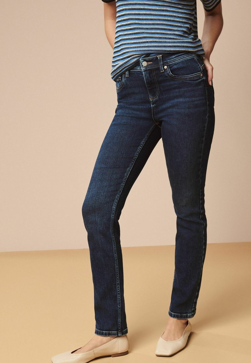 Mörkblå smala jeans med en slät tygtextur, femficksdesign och subtil sömnad, kombinerat med ljusa lägenheter.