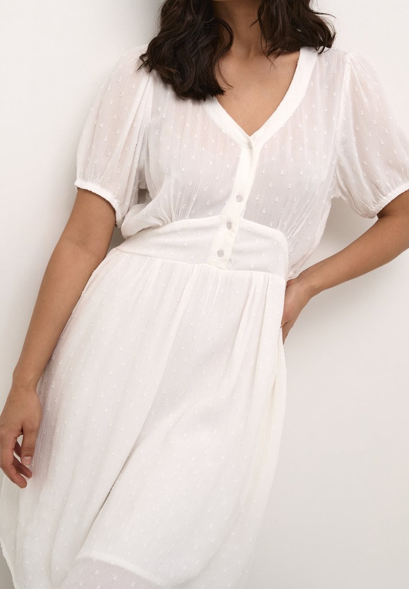 Femme portant une robe blanche texturée à manches courtes avec col en V, boutonnée à l'avant et taille cintrée, posant devant un fond uni.