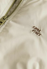 Beige lättviktsjacka med en framträdande dragkedja och broderad text "COOL VIBES" i brunt. Slät textur med minimala designdetaljer.
