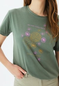 Camiseta de algodón color verde olivo con cuello redondo, manga corta y un gráfico colorido que presenta dientes de león y texto en rosa y dorado.