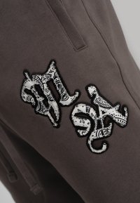 Pantalones gris oscuro con un parche blanco contorneado en negro que presenta un patrón de paisley negro formando letras estilizadas cerca del bolsillo de la cadera.