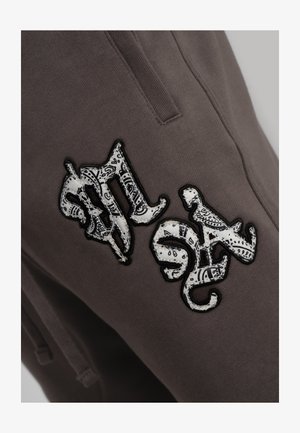 Donkergrijze broek met een zwartomlijnde witte patch waarop een zwart paisleypatroon staat dat gestileerde letters vormt nabij de heupzak.