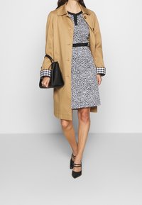 Tan trenchcoat över en svartvit prickig klänning med midjebälte; rutiga ärmuppslag. Svart handväska och svarta klackar.