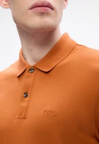 Vīrietis, tērpies oranžā polo krekliņā ar divām melnām pogām un vārdu "BOSS" izšūtu uz krūtīm.