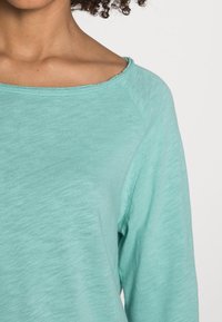 Helles türkisfarbenes Langarmshirt aus weichem Stoff mit weitem Ausschnitt und sichtbaren Nähten an Saum und Schultern.