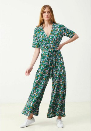 Kvinde med lyst hår iført en grøn blomsterprintet slå-om-dragt med korte ærmer og hvide sneakers, stående foran en ensfarvet baggrund.