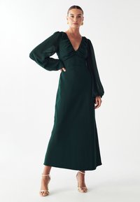 Dlhé zelené šaty s hlbokým výstrihom do V, volánikovým corps a splývavými priehľadnými rukávmi. Hladká textúra; v kombinácii s strapcovými zlatými podpätkami.