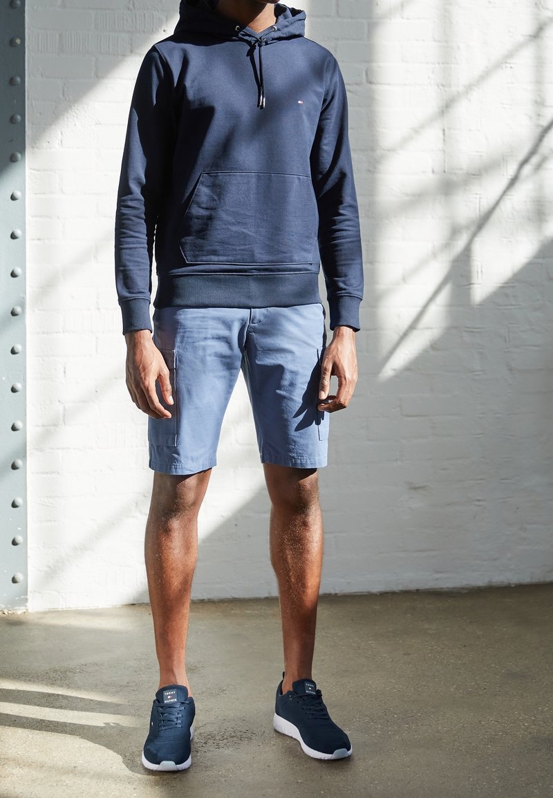 Navyblauer Hoodie mit Fronttasche, hellblaue Cargo-Shorts und navyblaue Sportschuhe mit weißen Sohlen. Glatte Textur und lässiges Design.