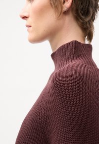 Maglione a collo alto in maglia bordeaux con texture a coste, caratterizzato da un colletto aderente e dettagli sulle spalle, mostrato di lato.