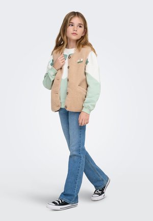 Beige fleecevest med knapper over en lysegrøn og hvid sweatshirt, kombineret med blå vide jeans og sorte sneakers.