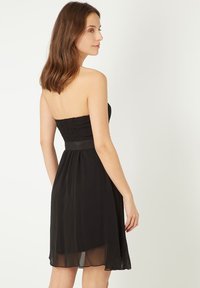 Vestido negro sin tirantes con un cuerpo ajustado de smock, falda acampanada y una capa translúcida desde la cintura hacia abajo, que muestra una textura suave.