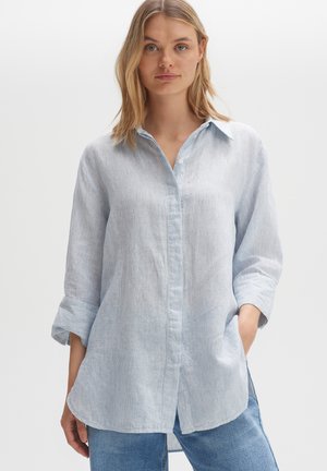 Woman with blonde hair wearing light blue striped long-sleeve shirt with rolled cuffs and blue jeans, standing with hands in pockets.