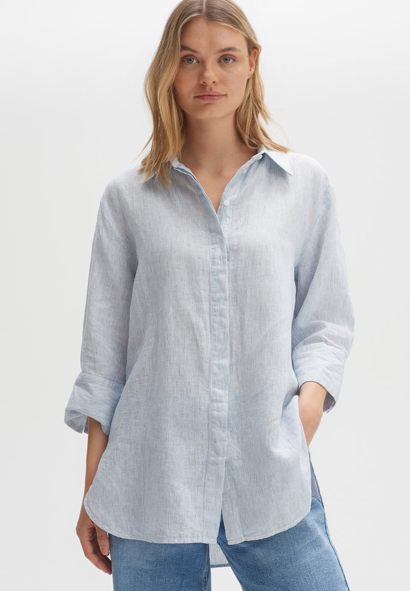 Woman with blonde hair wearing light blue striped long-sleeve shirt with rolled cuffs and blue jeans, standing with hands in pockets.