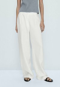 Des pantalons légers à jambes larges en tissu crème avec une texture douce, dotés d'une taille simple et de poches latérales, associés à un haut gris ajusté.