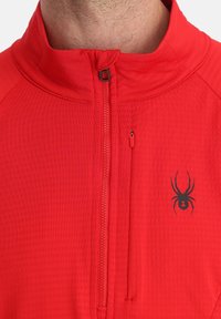 Rote Zip-Jacke mit hohem Kragen, aus strukturiertem Stoff und einem schwarzen Spinnenlogo auf der Brust, einschließlich einer Reißverschlusstasche.