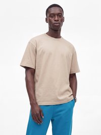 Junger Mann trägt ein beiges kurzärmeliges T-Shirt und blaue Hose und steht vor einem schlichten weißen Hintergrund.
