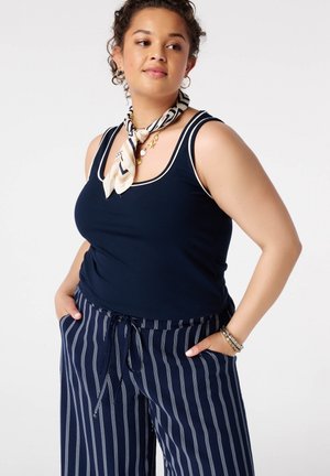 Vrouw die een marineblauw mouwloos topje, gestreepte marineblauwe en witte broek, een gestreepte halsdoek en gelaagde gouden kettingen met armbanden draagt, poseert met handen in de zakken.