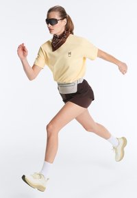 Femme courant portant un t-shirt jaune, un short marron, des baskets blanches, un bandana marron, des lunettes de soleil noires et une sacoche grise à la taille sur fond blanc.