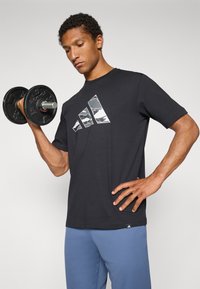 T-shirt en coton noir avec trois motifs de triangles en camouflage blanc et gris à l'avant ; associé à un pantalon de sport bleu, tenant un haltère.