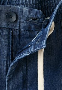 Pantalon en jean bleu foncé avec une fermeture éclair ouverte, deux boutons noirs et un cordon blanc visible à l'intérieur de la taille.