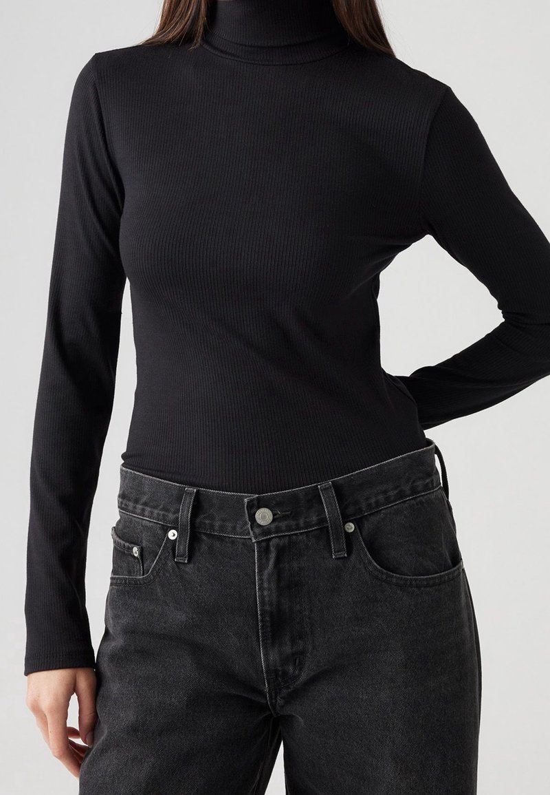 Un haut noir en maille côtelée à col roulé et manches longues associé à un jean en denim foncé avec fermeture à bouton et poches avant. Détail de tissu texturé visible.
